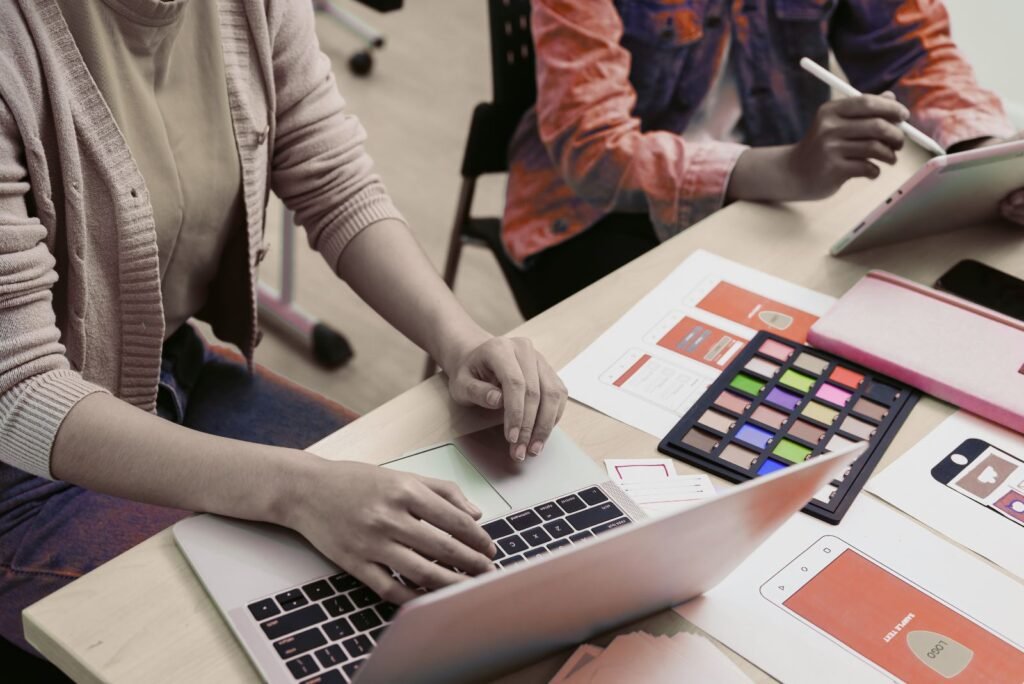 Two UI/UX designers collaborate on digital interfaces, using a laptop, tablet, wireframes, and a color palette.
