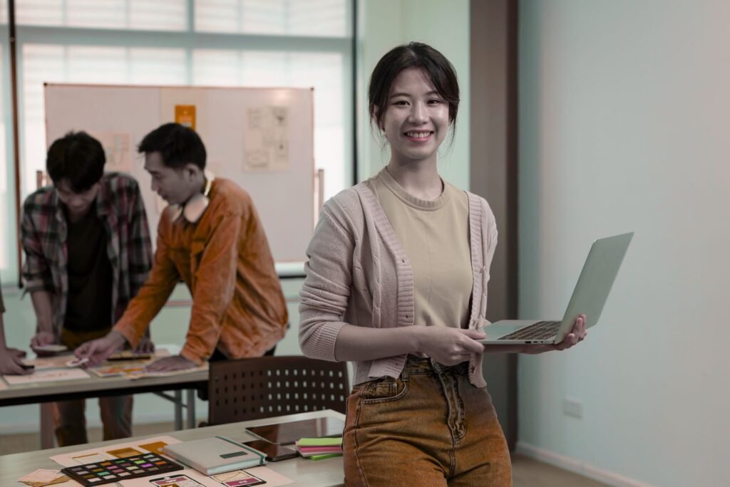 Smiling UI/UX expert holds laptop while team members collaborate on innovative digital interface designs.
