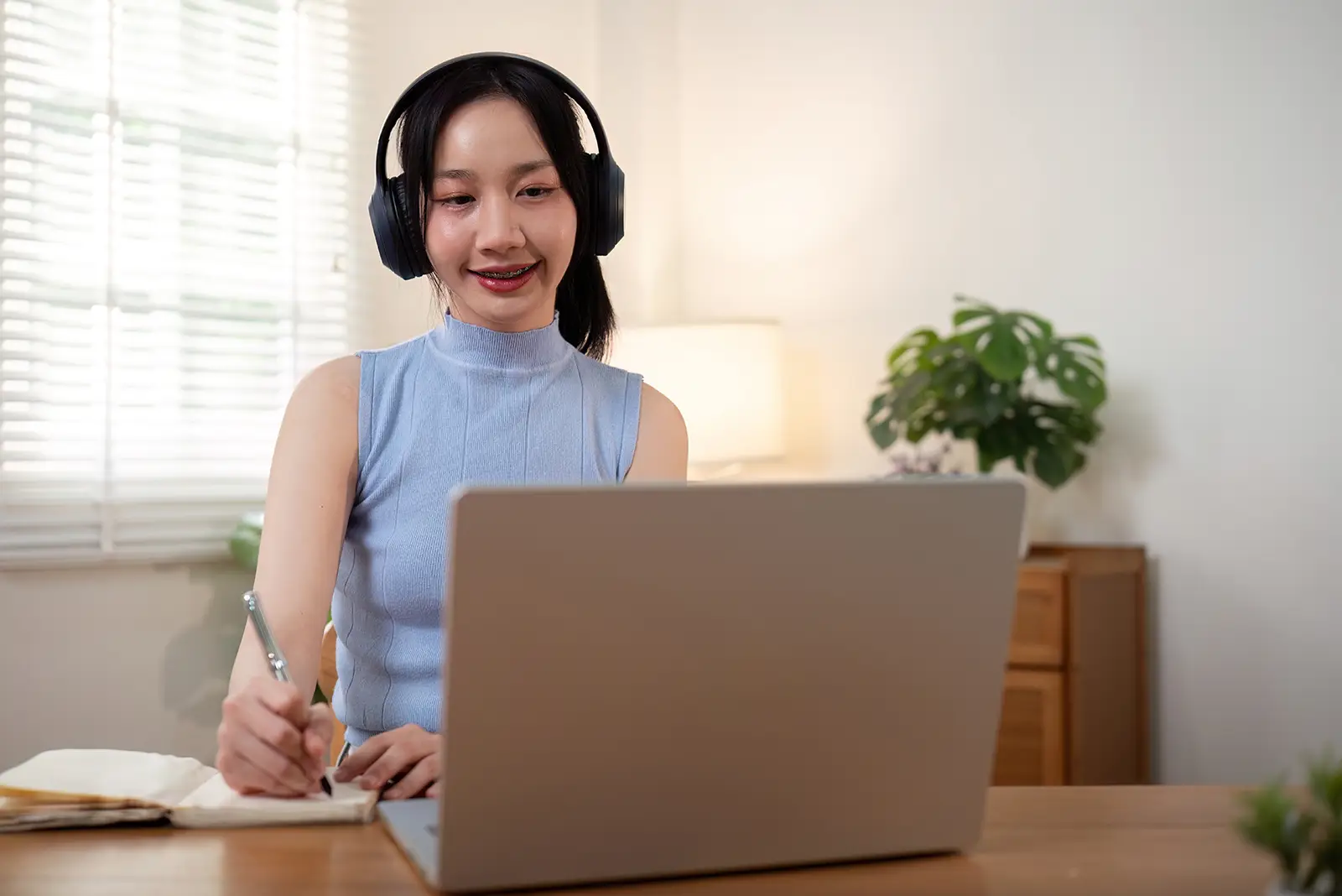 Smiling student wearing headphones, actively learning a language online, taking notes from a laptop for ALA Language School.