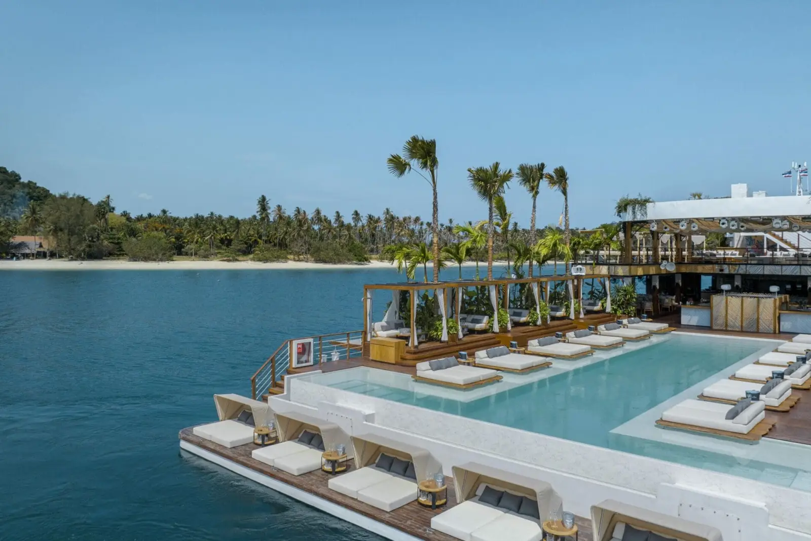 Yona Beach Club, a floating club in Phuket, with a large pool, sun loungers, palm trees, and a tropical island backdrop.
